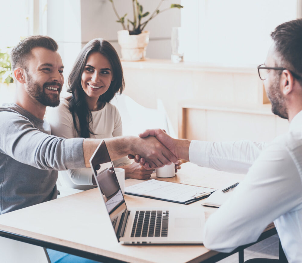 Good deal! Cheerful young man bonding to his wife while shaking hand to man sitting in front of him at the desk